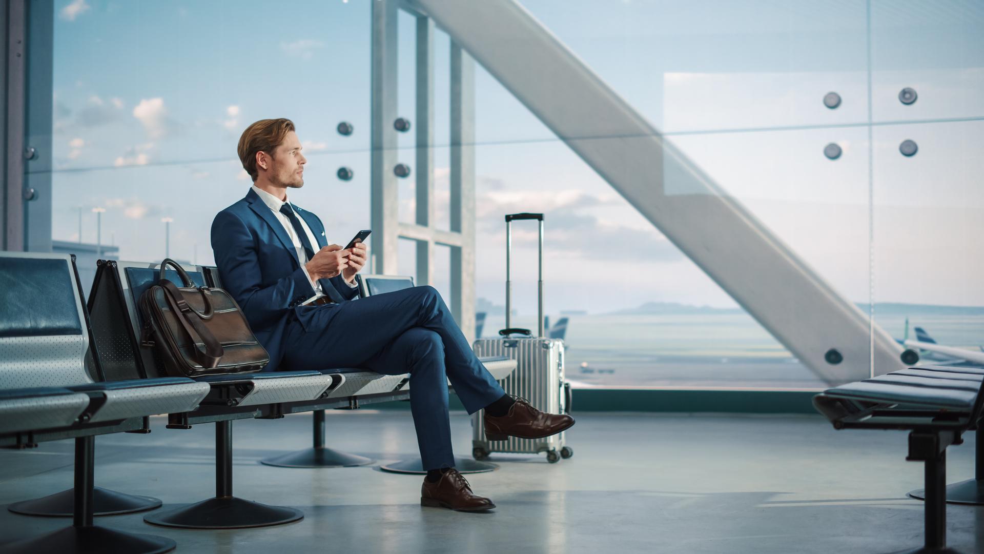 Airport Terminal: Businessman Uses Smartphone, Waiting for a Flight, Doing e-Business, Sending e-Commerce Data. Traveling Man Remote Work Online on Mobile Phone in Boarding Lounge of Airline Hub